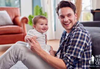 A father carrying his child while sitting on the ground beside a couch in a living room.